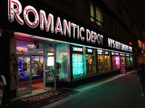 Exterior view of Romantic Depot store at night with neon signs promoting lingerie, toys, and over 100,000 items.