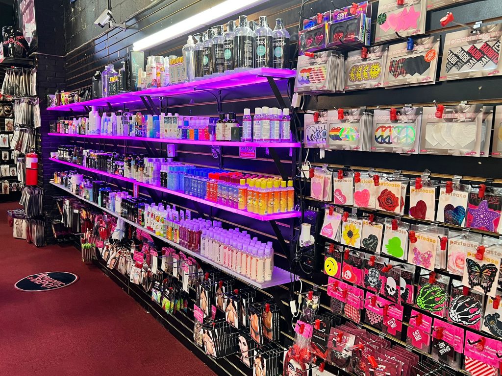 Romantic Depot store shelf with lubricants, toy cleaners, and colorful nipple pasties under purple lighting