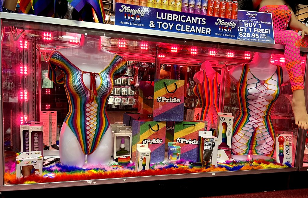 Romantic Depot Pride display with rainbow lingerie, LGBTQ+ gift bags, sex toys, and lubricant under pink lighting