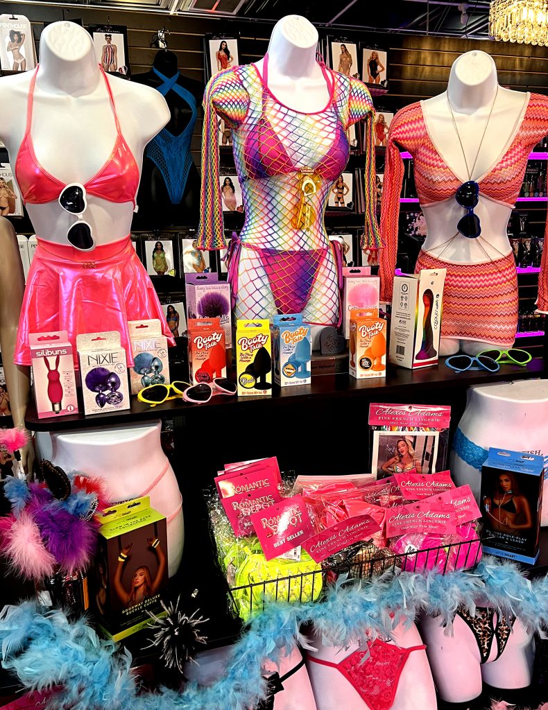 Romantic Depot display with rainbow fishnet lingerie, pink outfits, butt plugs, and adult novelty items in a vibrant Pride-themed setup