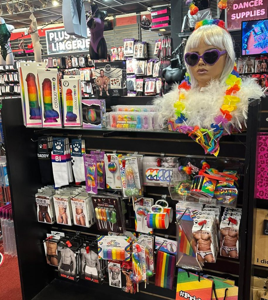 Display of Pride-themed adult products at Romantic Depot, featuring rainbow dildos, colorful underwear, Pride flags, accessories, and a mannequin in festive LGBTQ+ attire with heart-shaped sunglasses and feather boa.