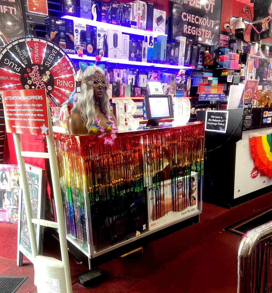 Romantic Depot checkout counter decorated with rainbow fringe and a mannequin in festive Pride gear, surrounded by sex toys and colorful signage celebrating LGBTQ+ inclusivity.