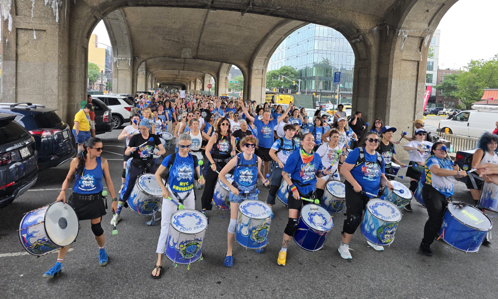 NYC drumline performing under a viaduct during SunnyPride parade, with a diverse group of drummers marching in celebration of LGBTQ+ pride.
